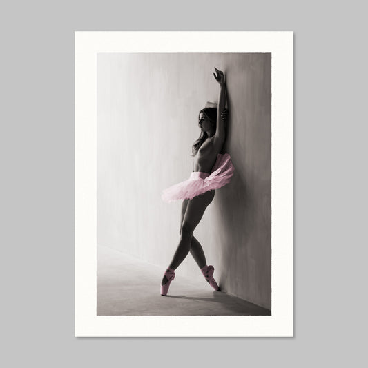 A vertical art print featuring a black and white photograph of a ballerina in a pink tutu and pointe shoes leaning against a wall, presented with a wide white border on a light gray background.